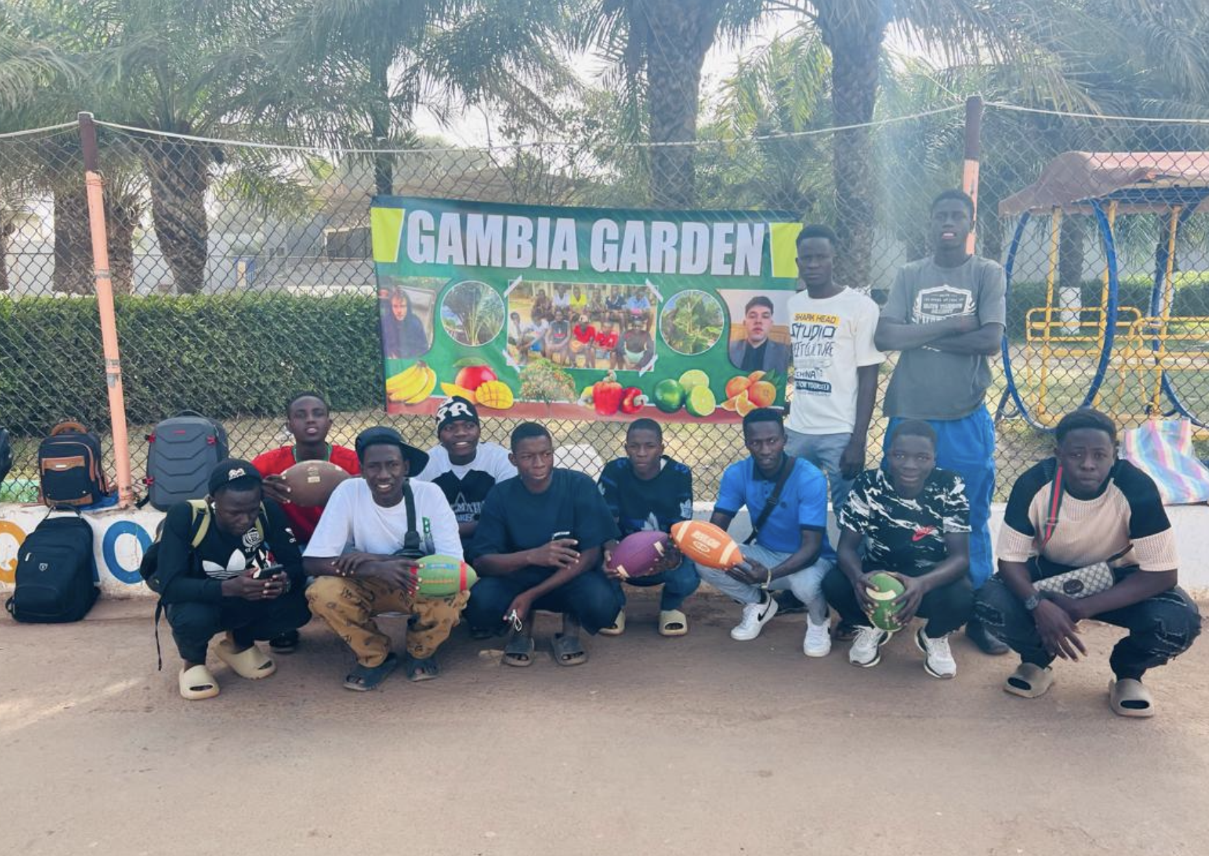 Flag Football participants holding footballs together in front of a Gambia Garden banner
